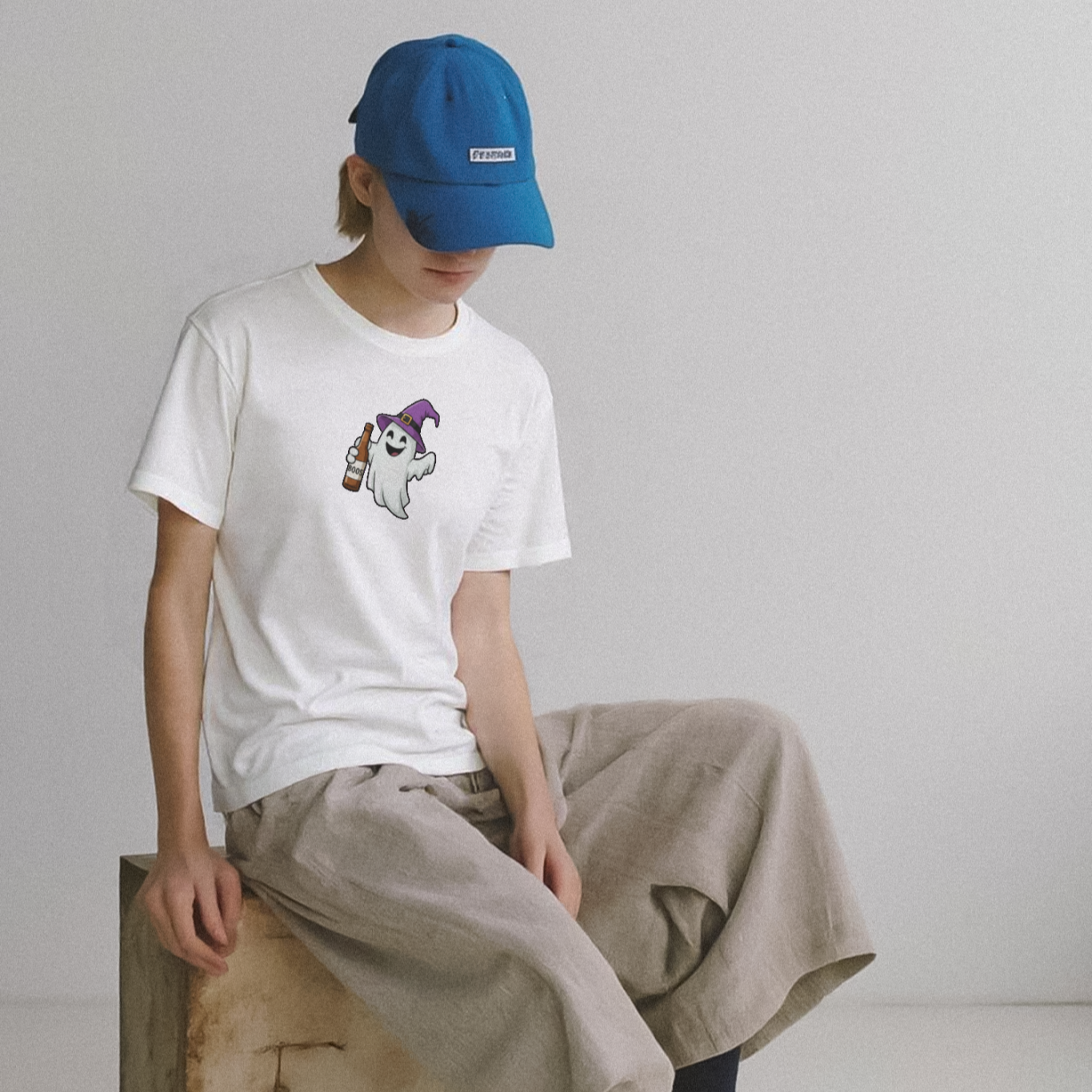 Person wearing a white t-shirt with a graphic, beige pants, and a blue cap sitting on a wooden block.