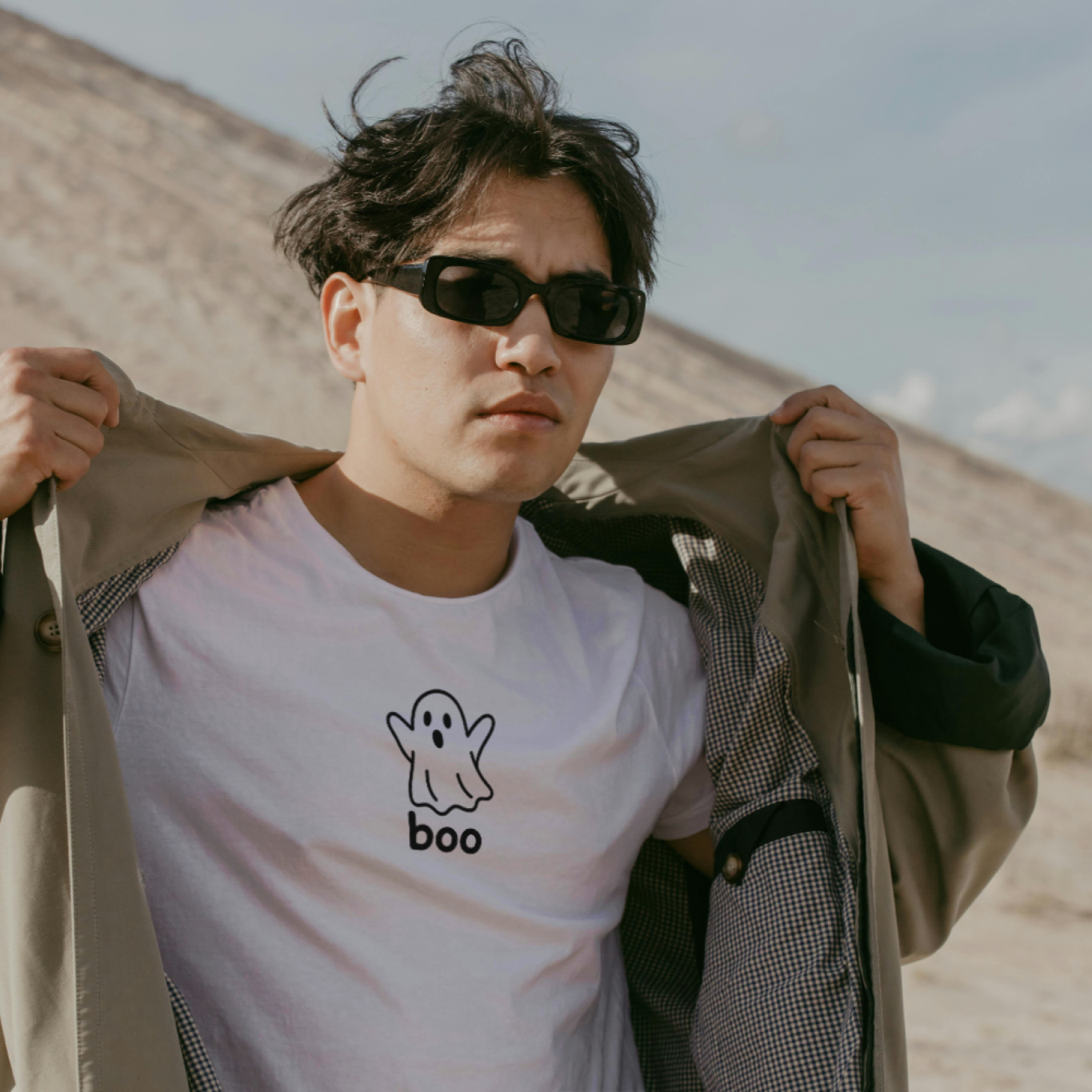 Man wearing sunglasses and a white t-shirt with a ghost design, standing in a desert-like setting.