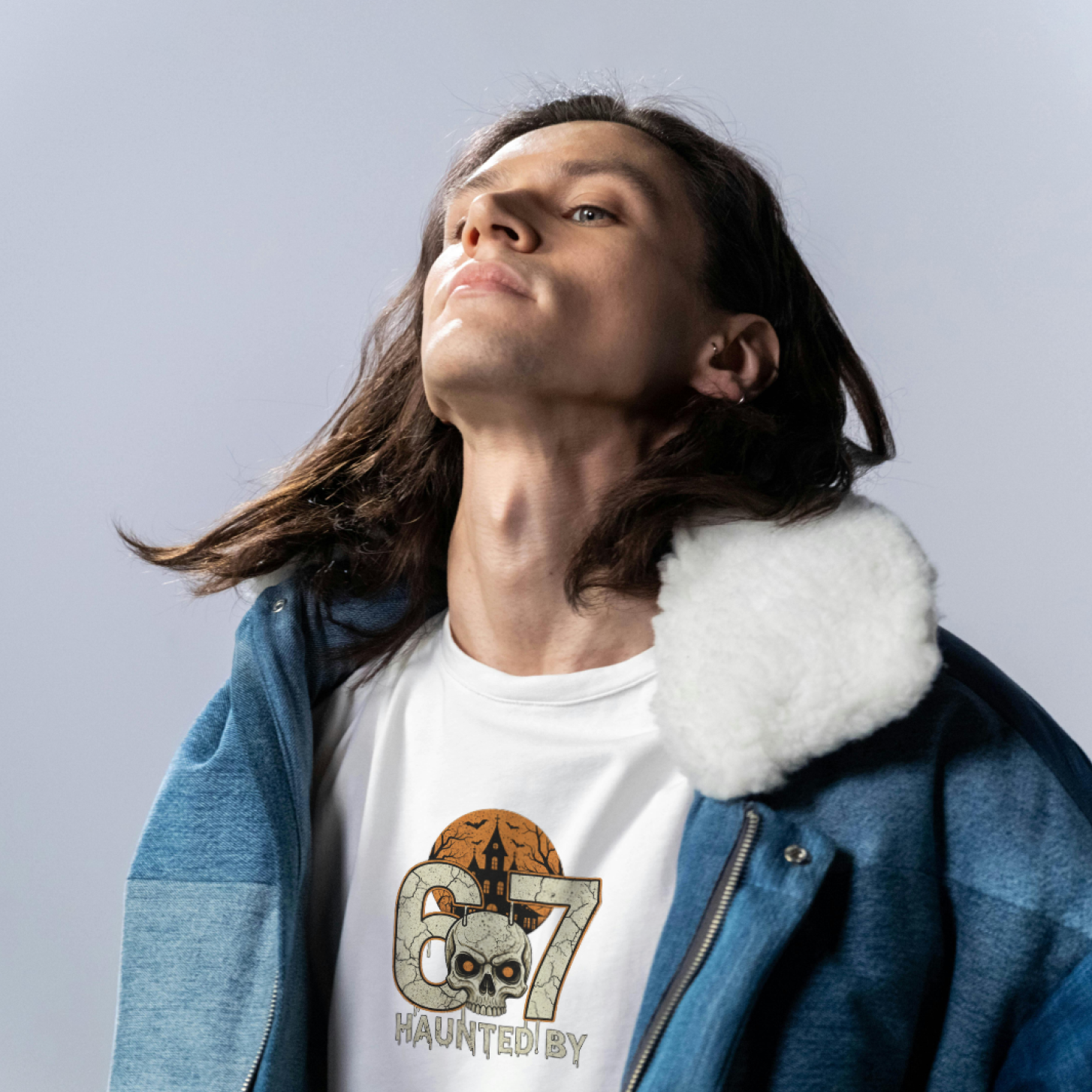 Person wearing a white t-shirt with a graphic design and a blue jacket with a fur-lined hood against a light gray background