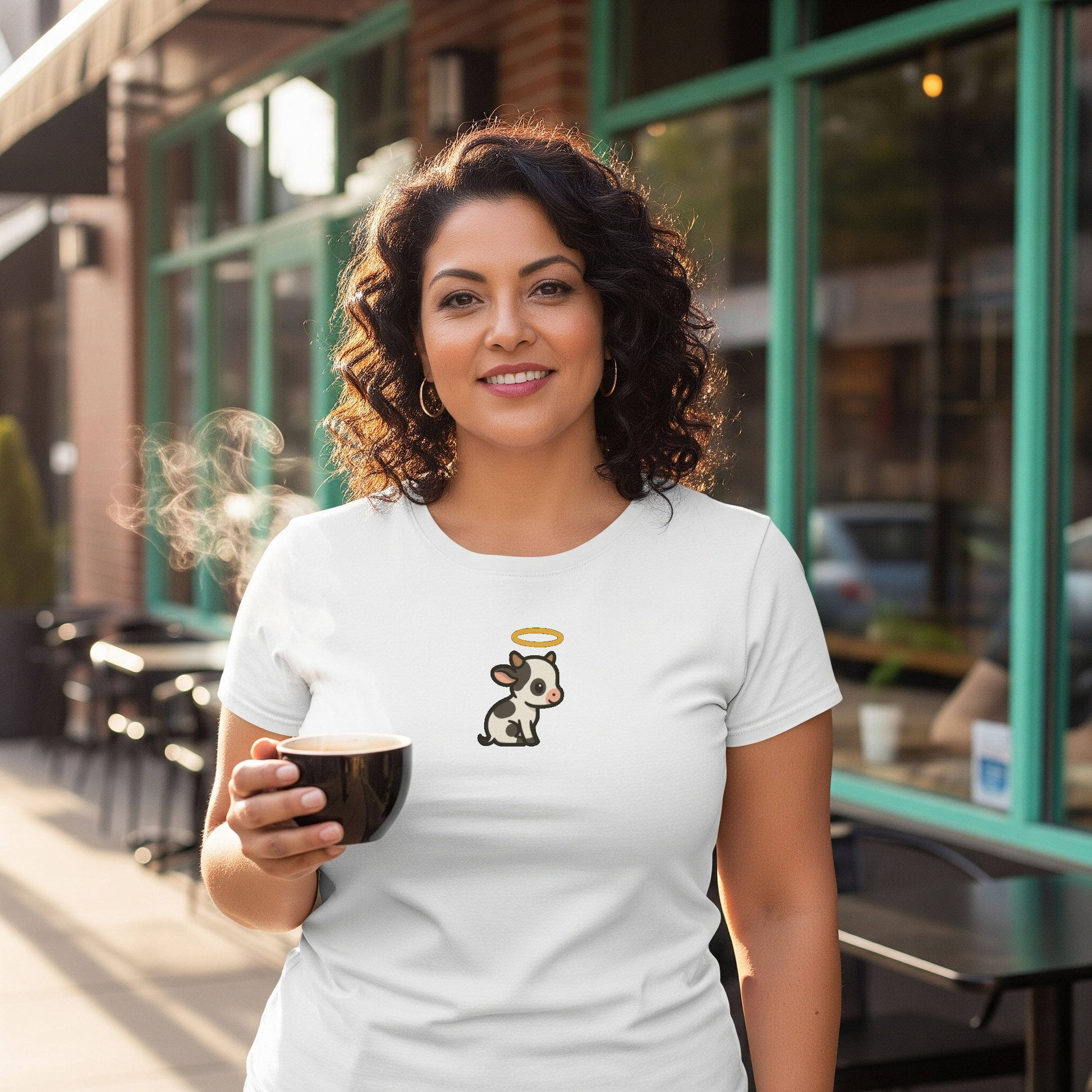 Woman holding a coffee cup outdoors, wearing a white t-shirt with a cow graphic and angel wings.
