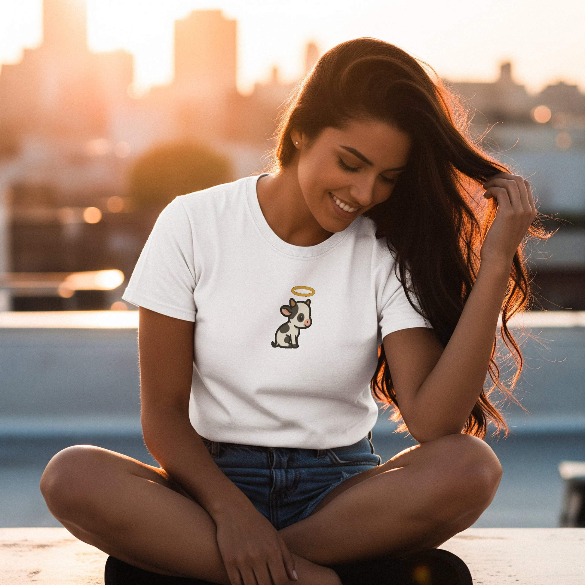 Woman sitting outdoors wearing a white t-shirt with a holy cow graphic, cityscape in the background.