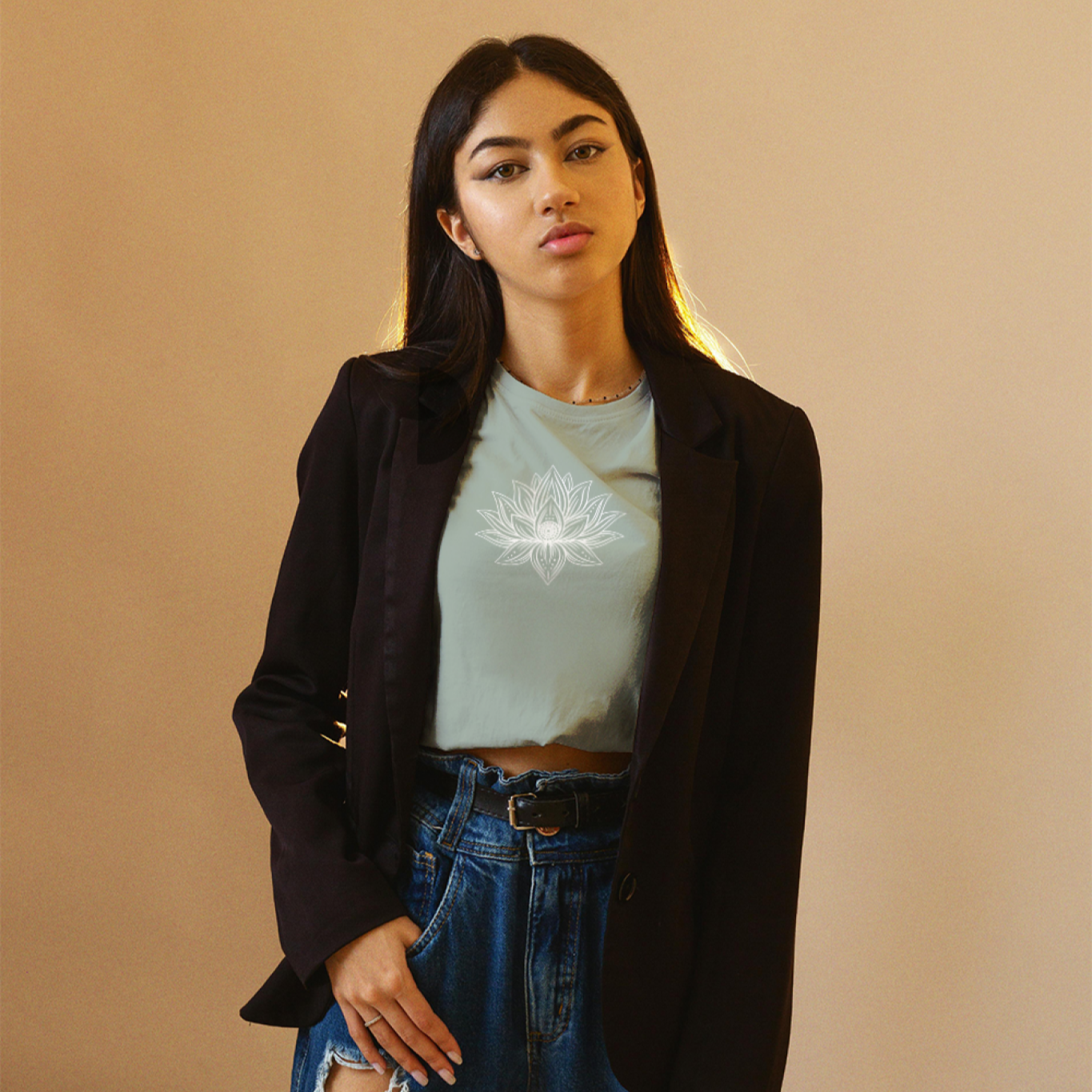 Woman wearing a black blazer, light blue shirt, and ripped jeans against a beige background