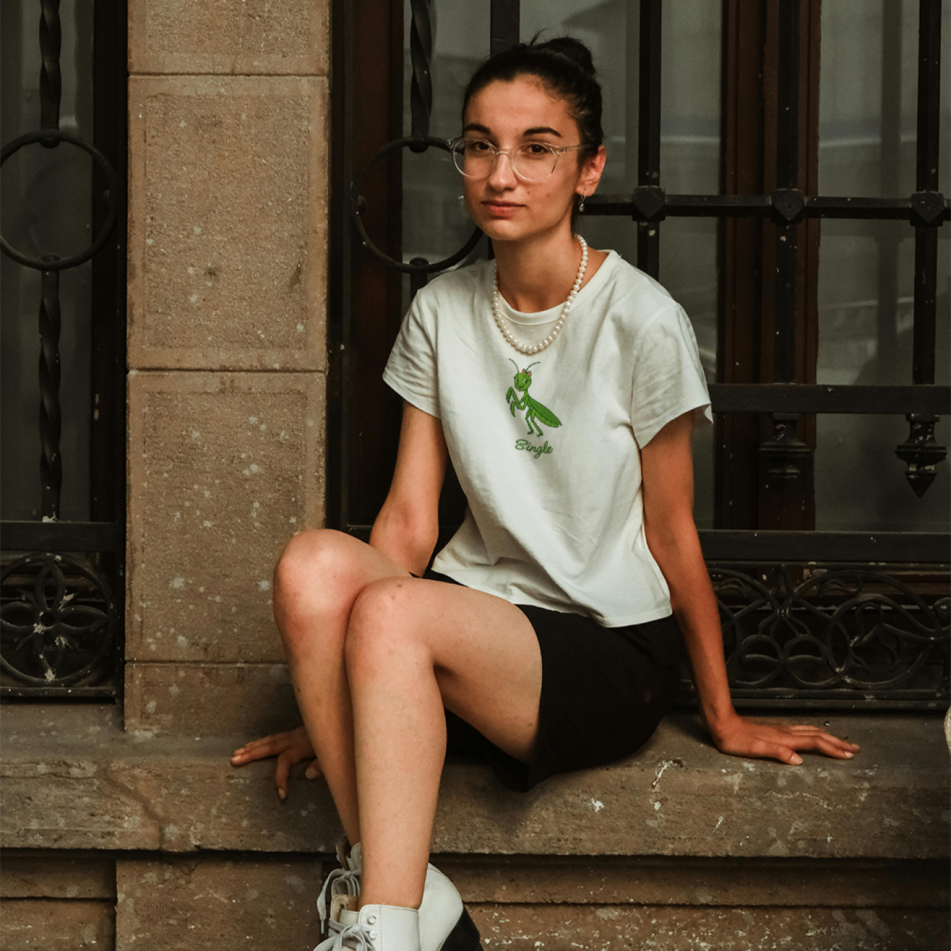 Person sitting on steps wearing a white t-shirt with a green graphic and black shorts, with roller skates on.