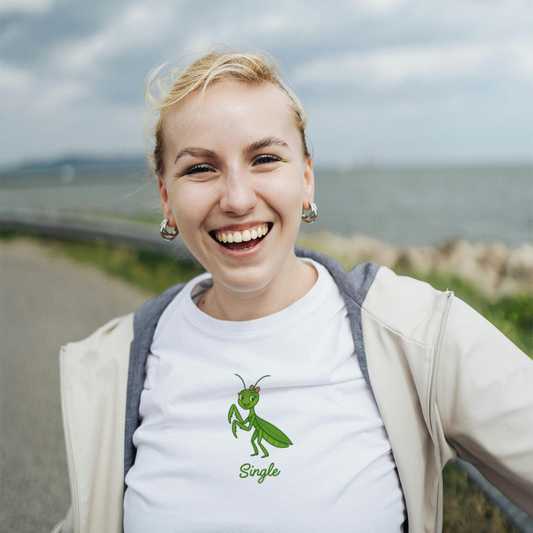 Woman smiling outdoors with a cloudy sky and water in the background, she is wearing a praying mantis shirt that says "Single"