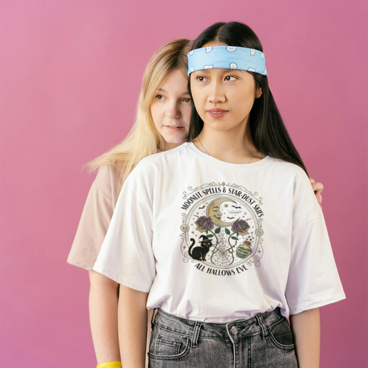 Two women standing against a pink background, one is wearing a white t-shirt with moonlit spells and star-dust skies shirt.