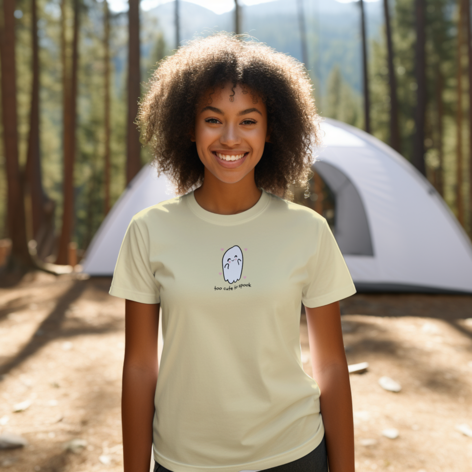 Person wearing a beige t-shirt with a graphic design in front of a tent in a forest setting
