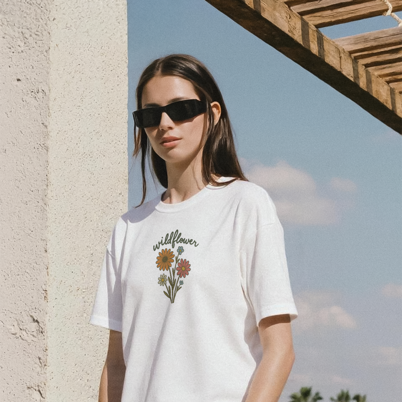 Woman wearing a white t-shirt with floral design and sunglasses, standing against a stone wall with a wooden pergola and palm trees in the background.